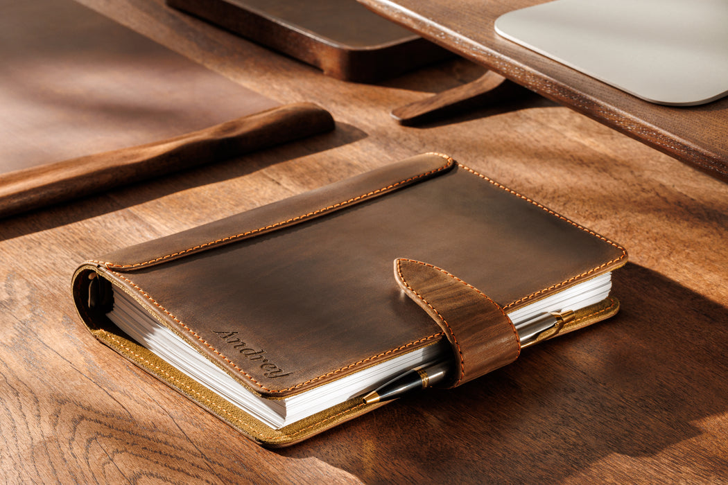 Top view of black Mureli notebook with engraved name on wooden surface near watch and pen