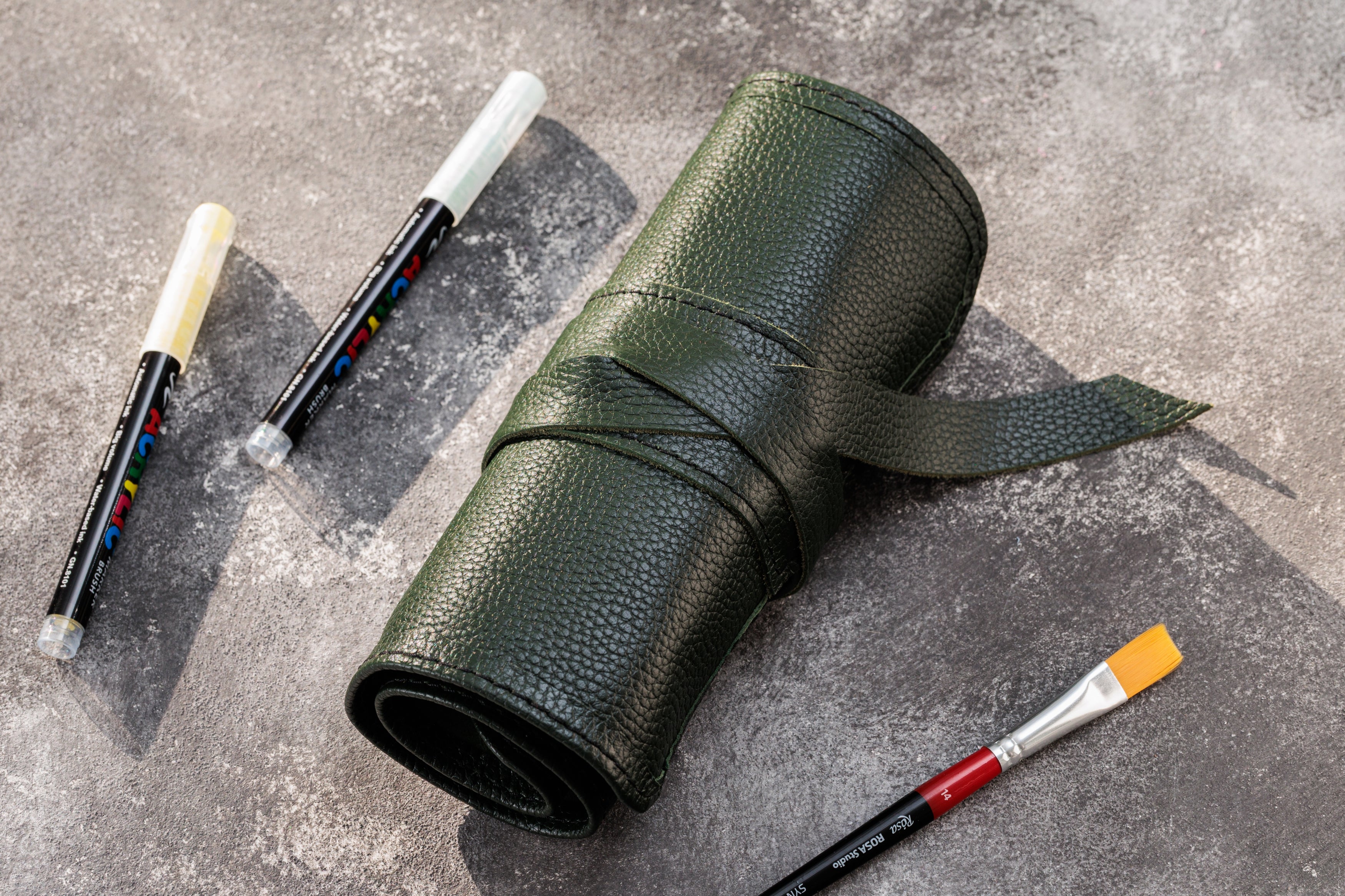Green leather pencil case with markers and paintbrushes on a gray surface