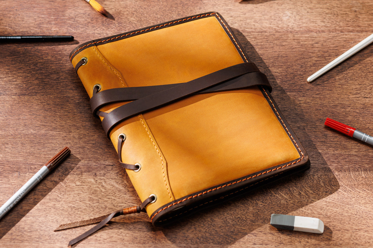 camel yellow leather notebook organizer with brown wrap strap on wooden table