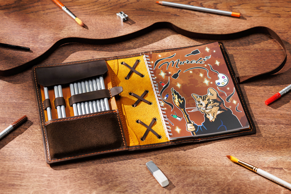 camel yellow and brown leather sketchbook organizer with pencils and spiral notebook on wooden table
