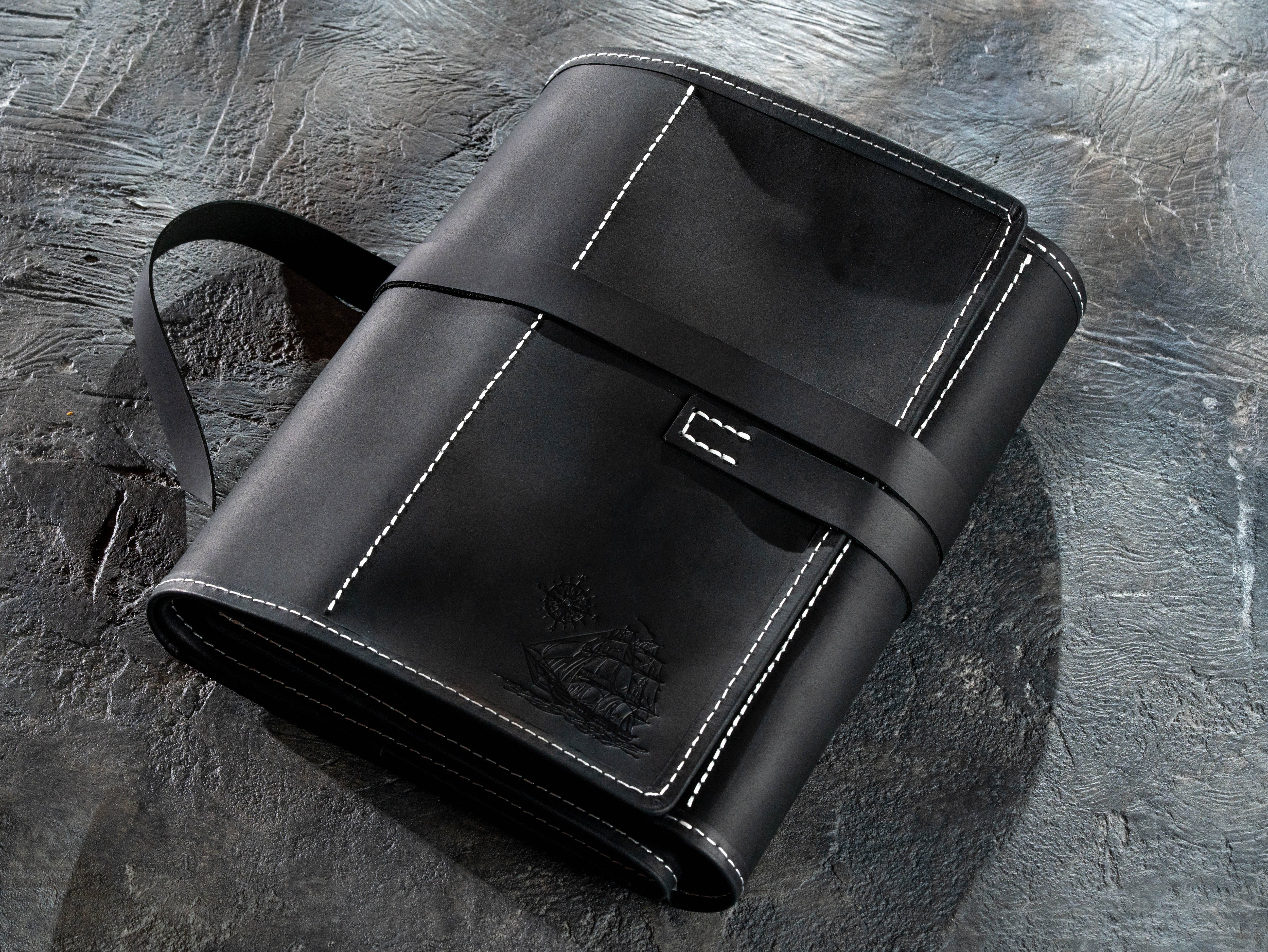 A black leather folio-style case with white contrast stitching, a wrap-around strap, and a small metal clasp, photographed on a textured dark gray surface.