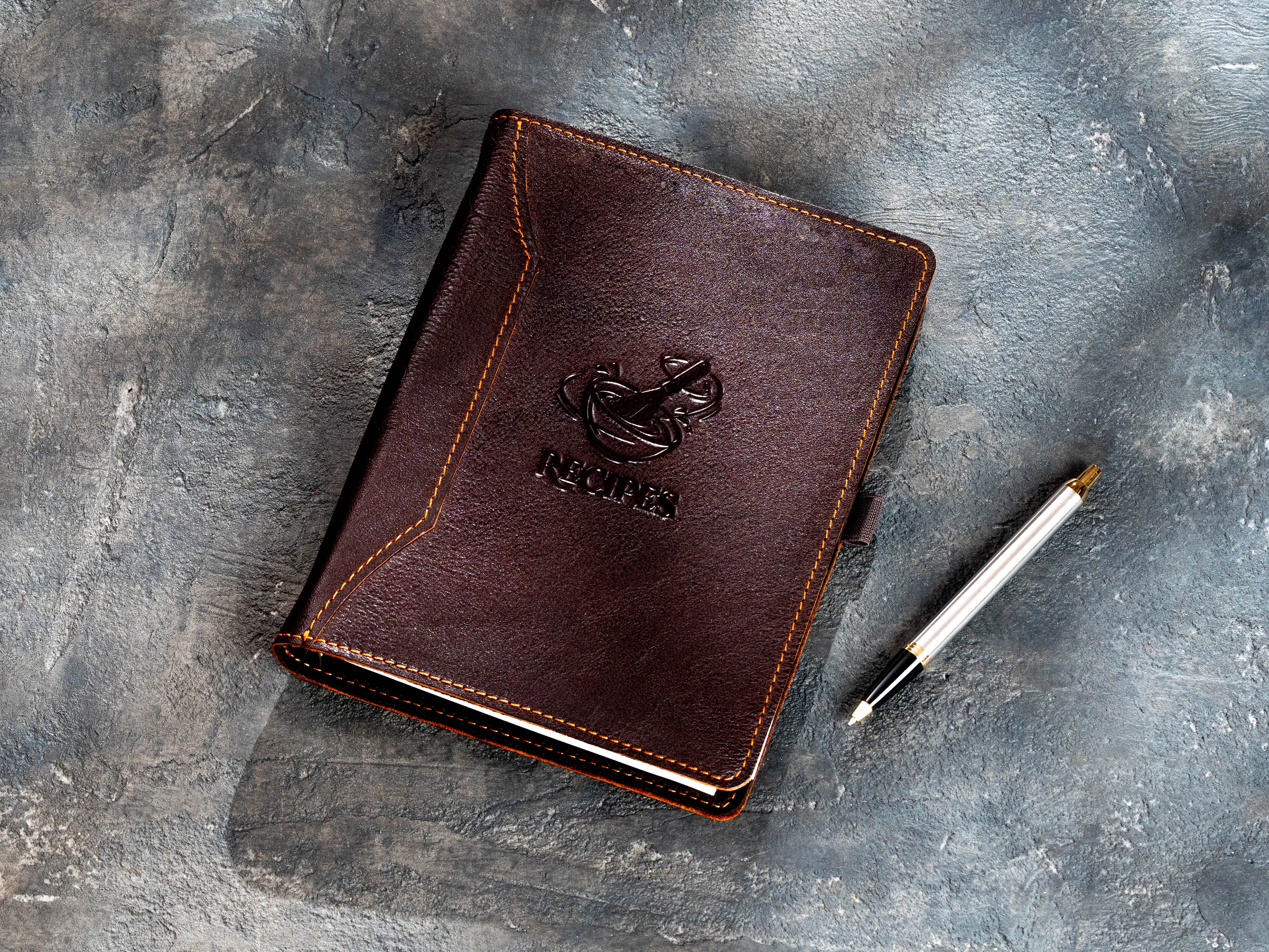 Dark brown leather recipe binder with embossed bowl and kitchen utensils design beside a pen