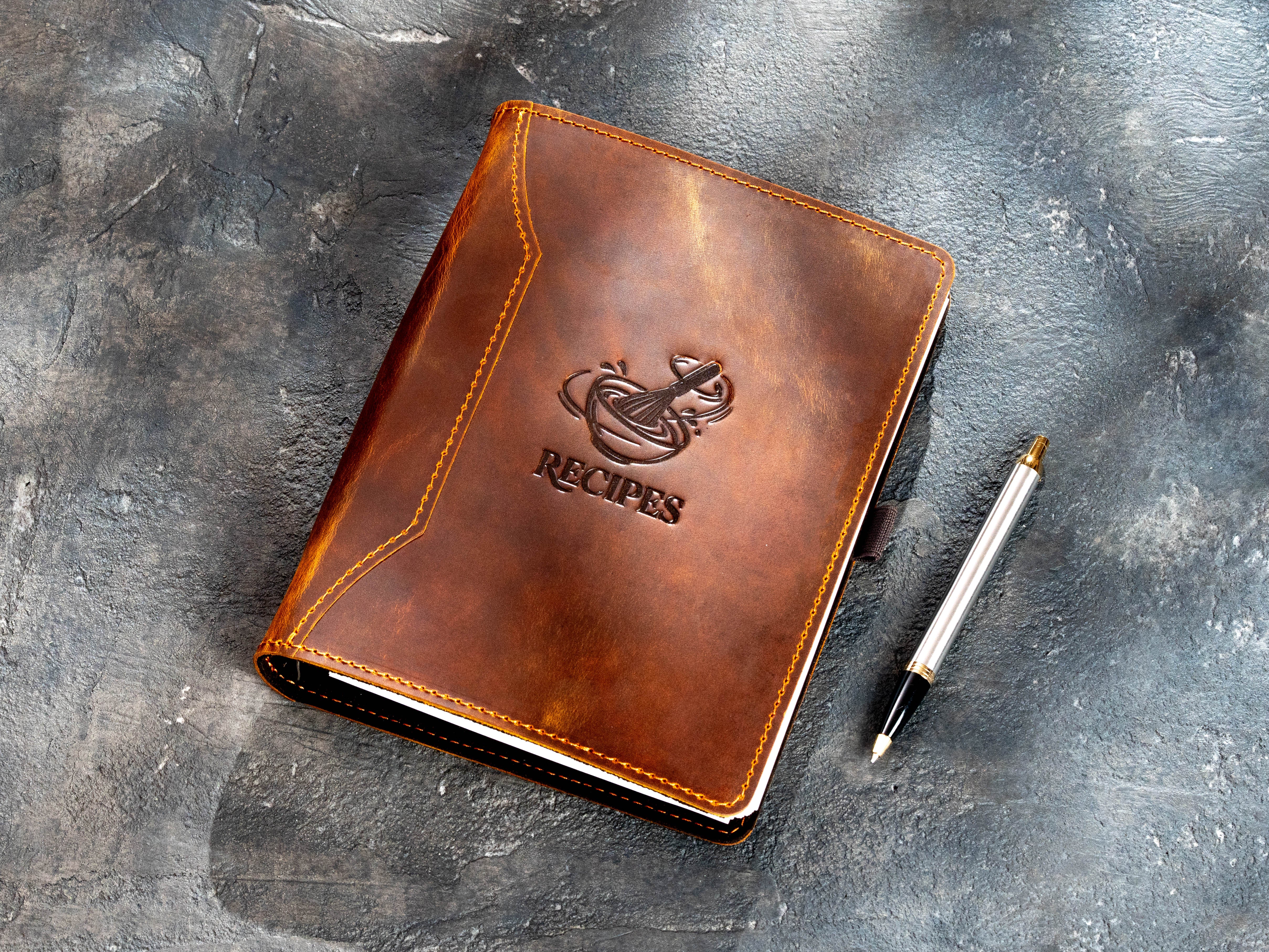 Chestnut leather recipe organizer with embossed bowl and kitchen utensils design beside a pen