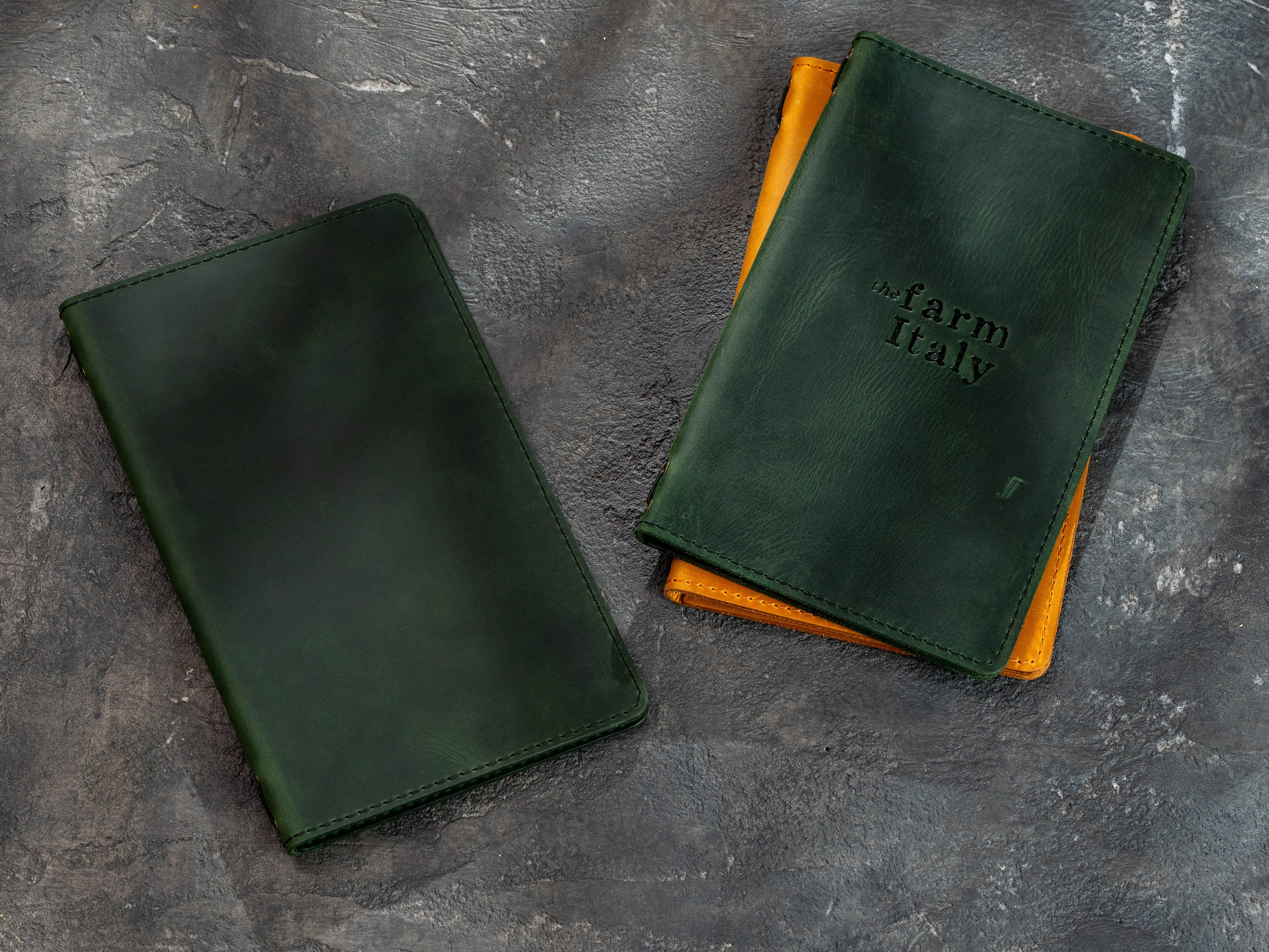 Two dark green leather folio-style cases on a textured gray surface, with one case standing upright and the other lying flat. The upright case has 'farm italy' embossed on the front and is stacked on top of a yellow-orange item.