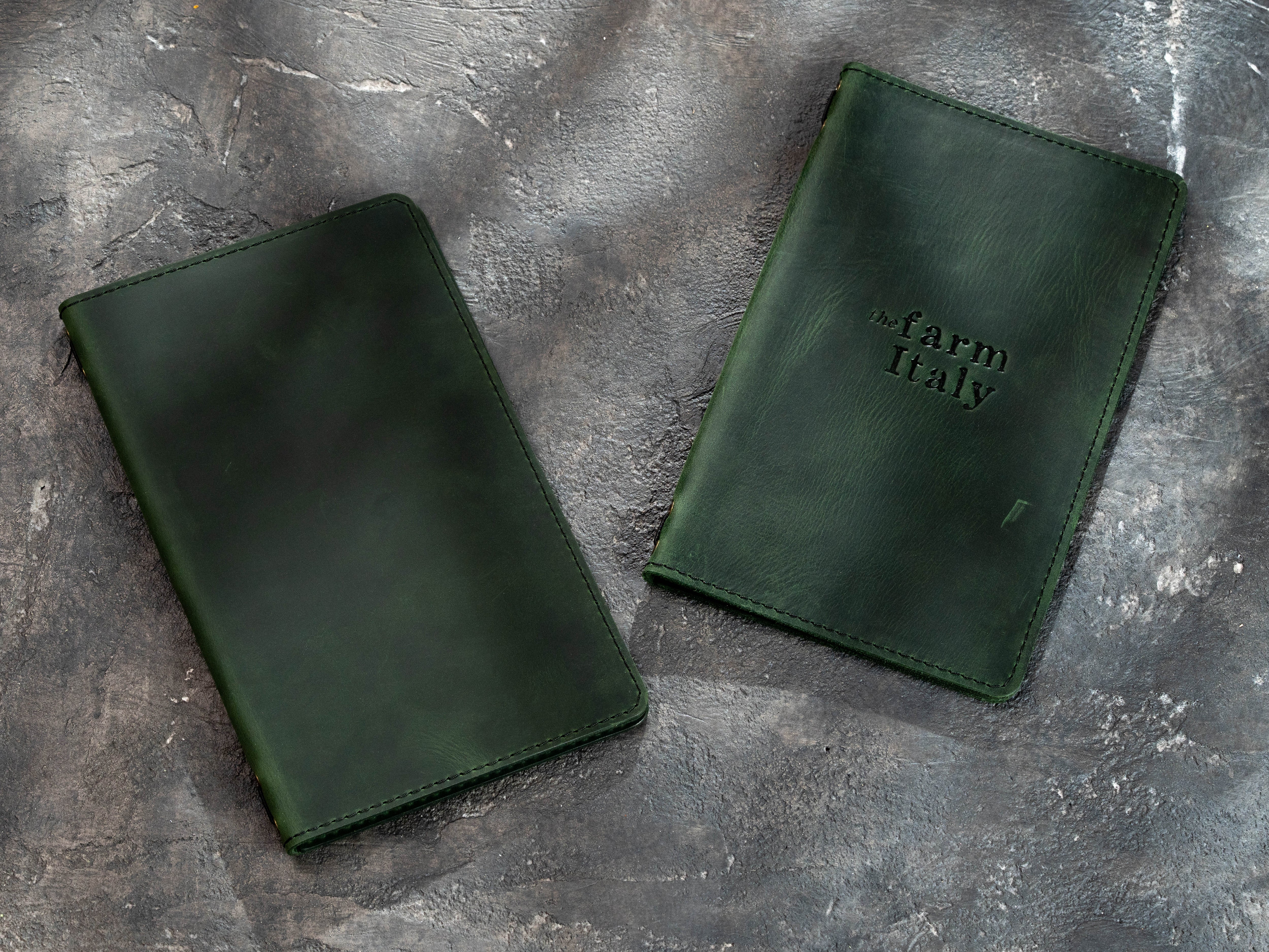 Two dark green leather folios or wallets displayed on a textured gray stone surface. One folio is plain, and the other features an embossed 'farm Italy' logo.