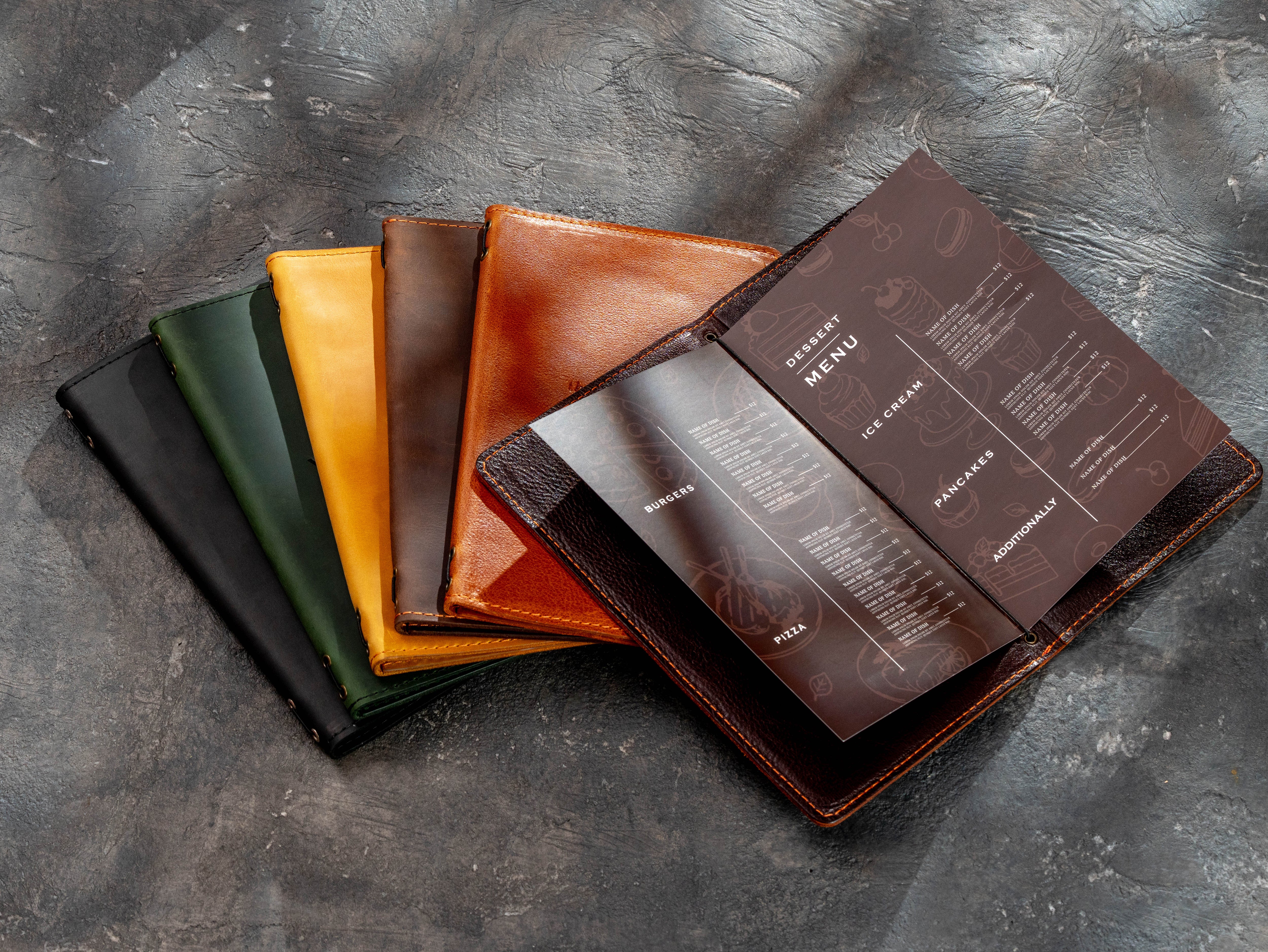 A set of leather menu folios or booklets in multiple colors, including black, green, yellow, brown, and reddish-brown, arranged on a textured dark gray surface. One reddish-brown folio is open, displaying printed menu pages.