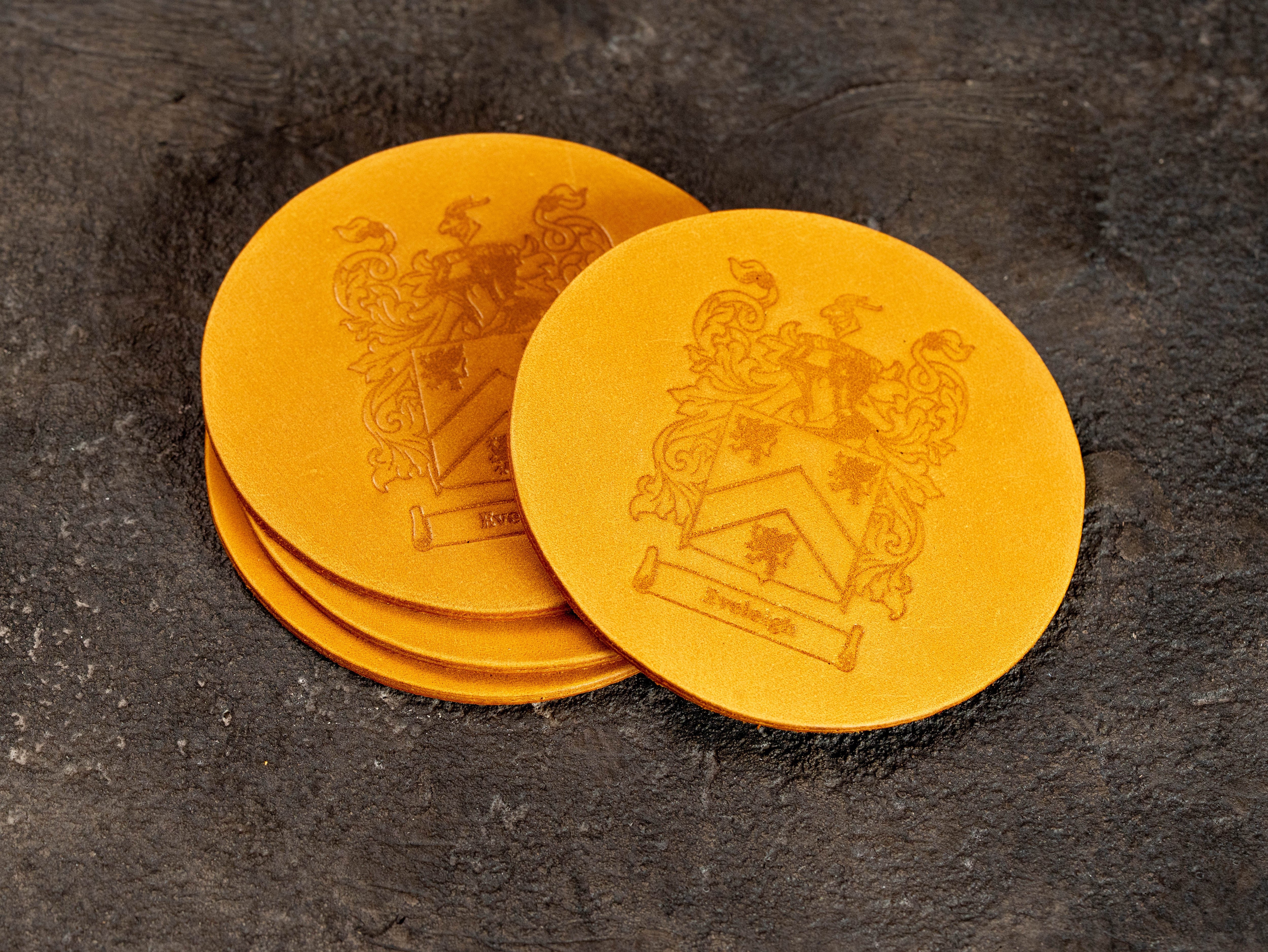 Custom leather drink coasters in wheat yellow with detailed family crest and name, designed as part of a coordinated personalized leather set.