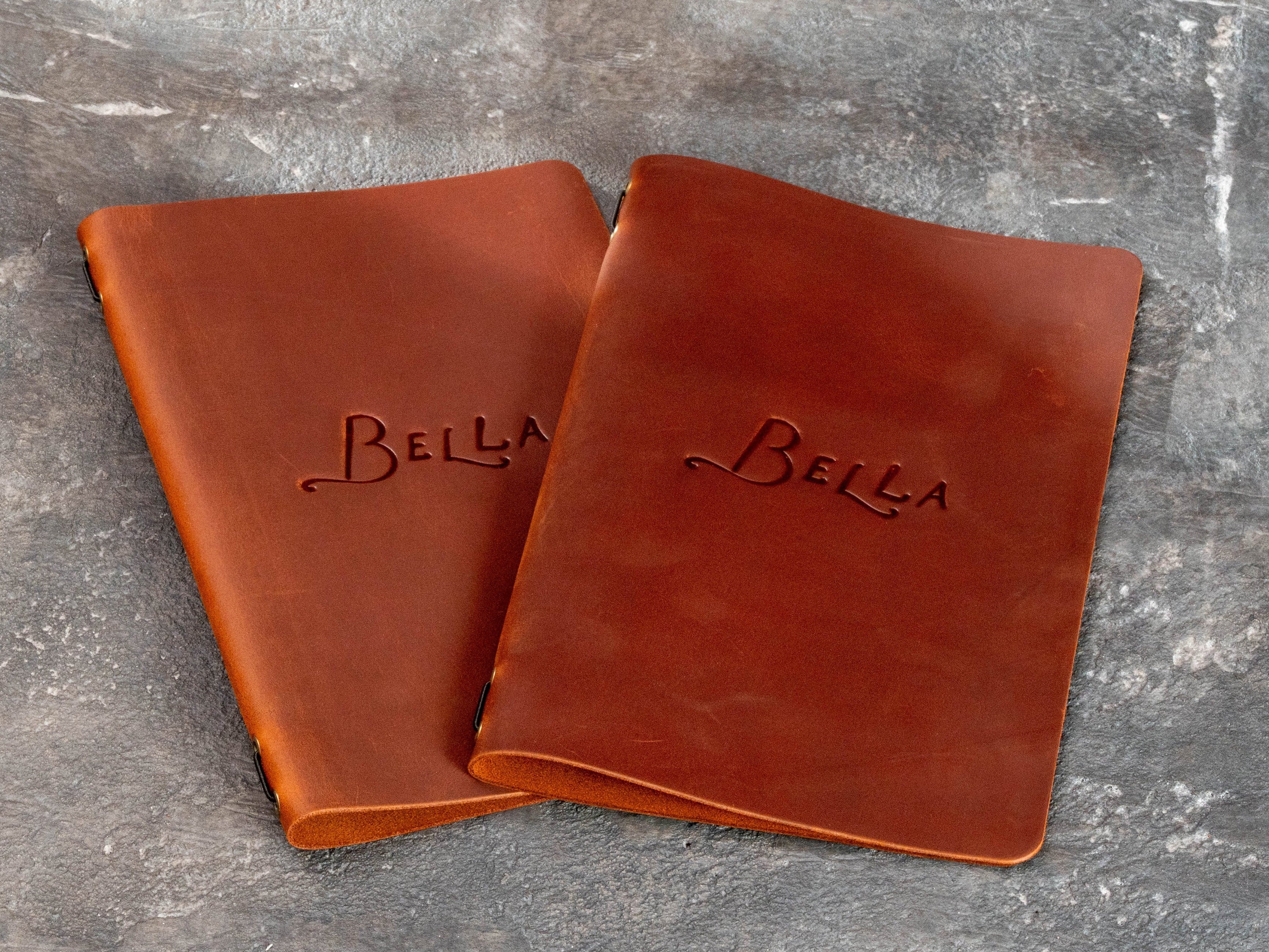 Two warm brown leather Bella menu covers with blind embossed restaurant branding and rounded corners