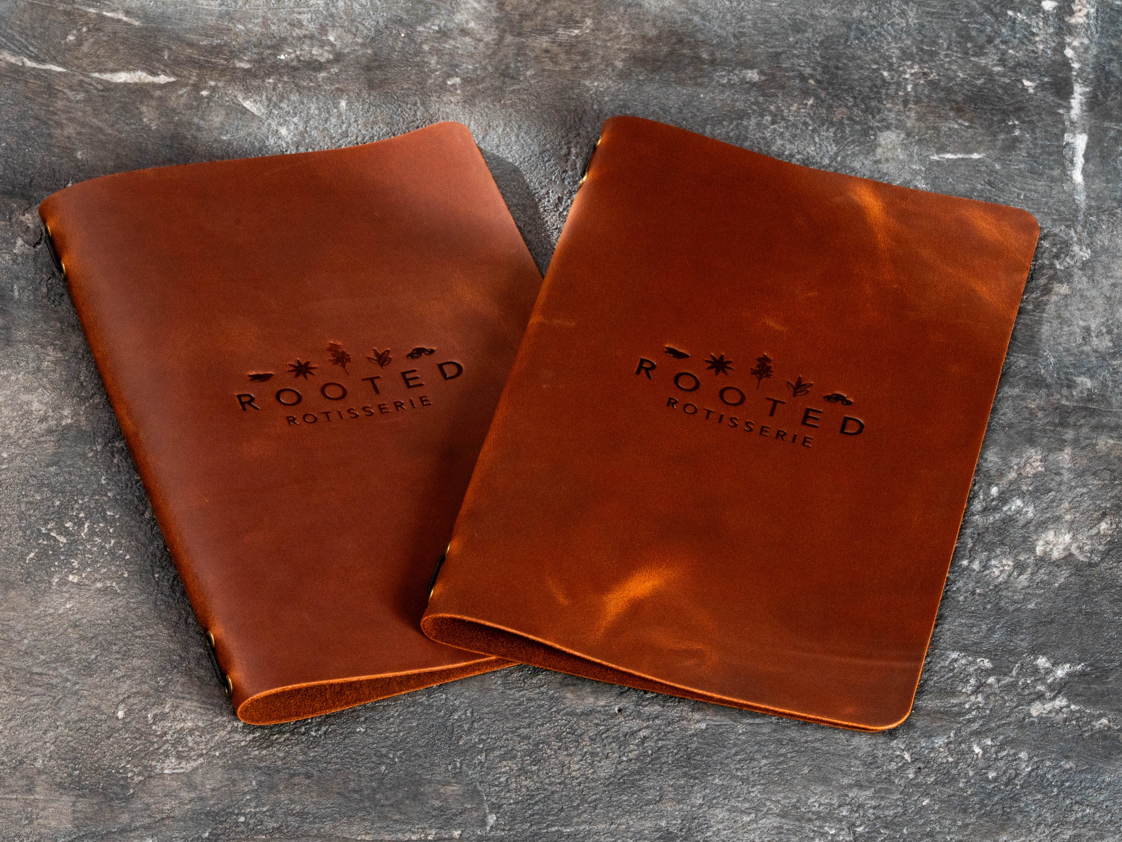 Two warm brown leather menu books with subtle blind embossed Rooted Rotisserie logos