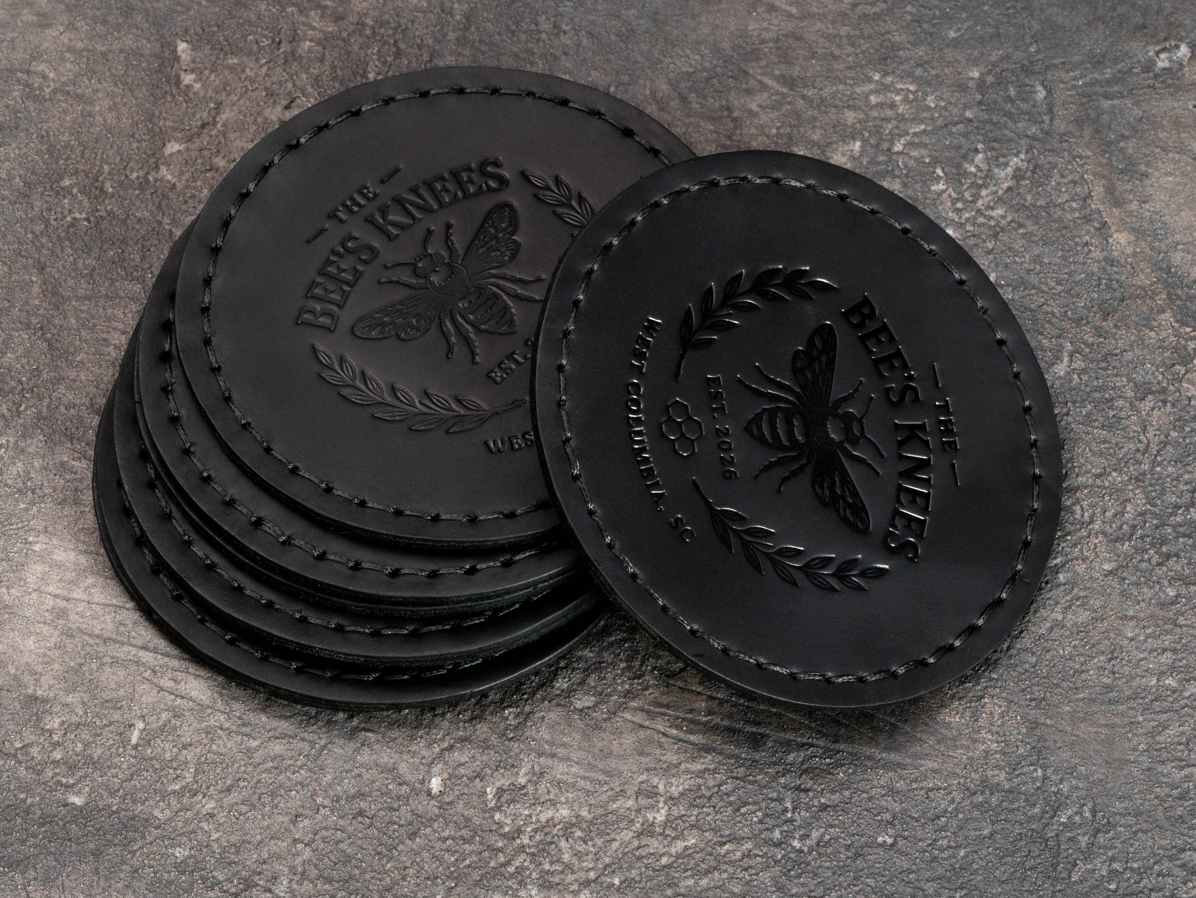 Stack of custom black leather founding member coasters with embossed bee emblem, club name, location and establishment year.