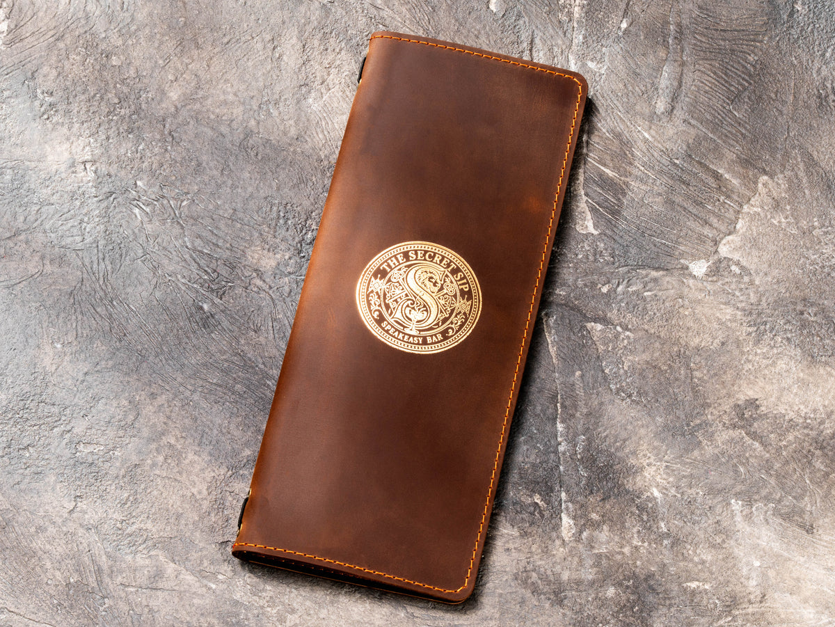 Brown leather cocktail menu cover with gold foil The Secret Sip logo for speakeasy bar and private event service