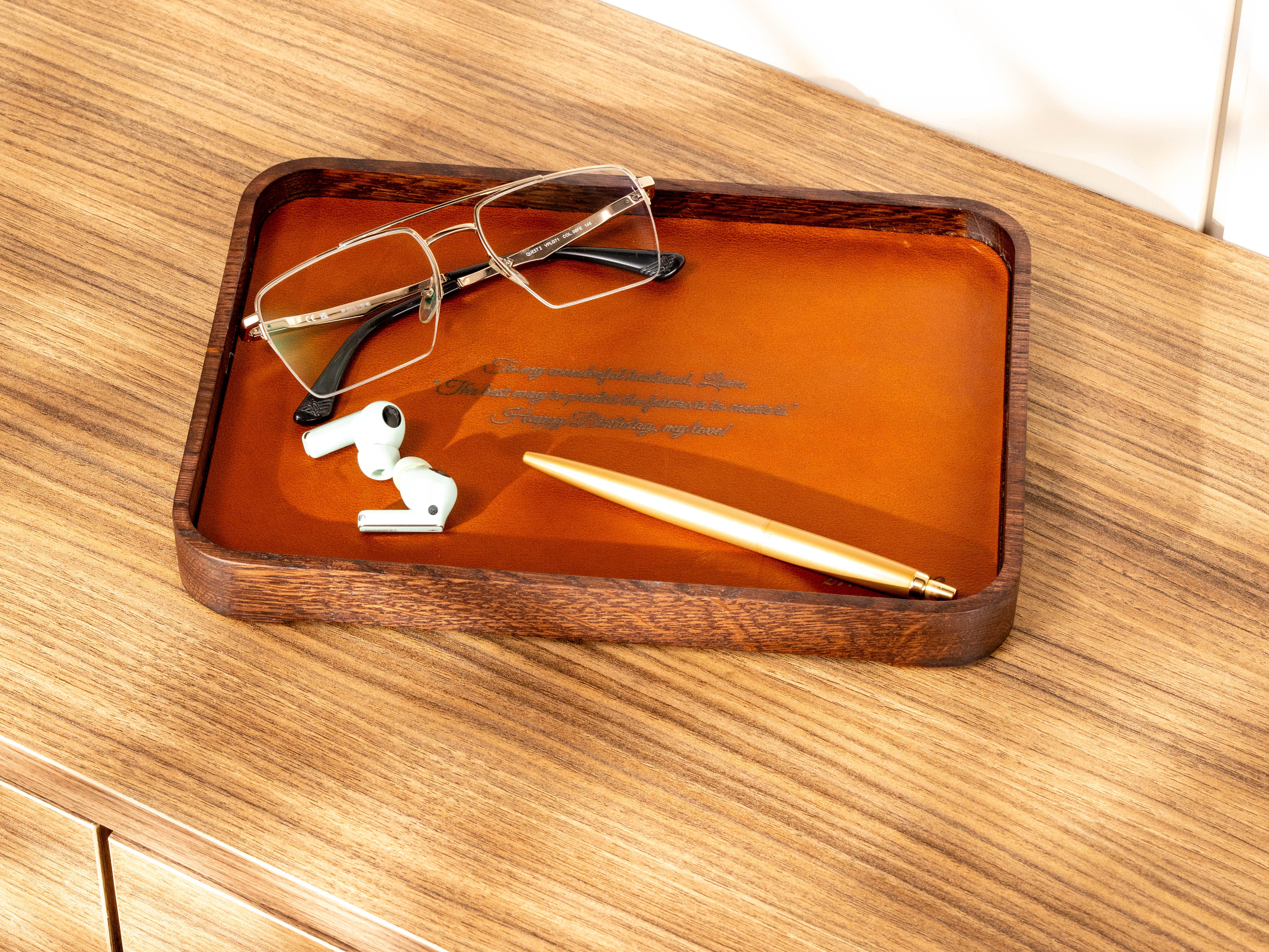 Oak and Camel leather catchall tray with personalized engraving, glasses, wireless earbuds and pen on a wooden dresser.