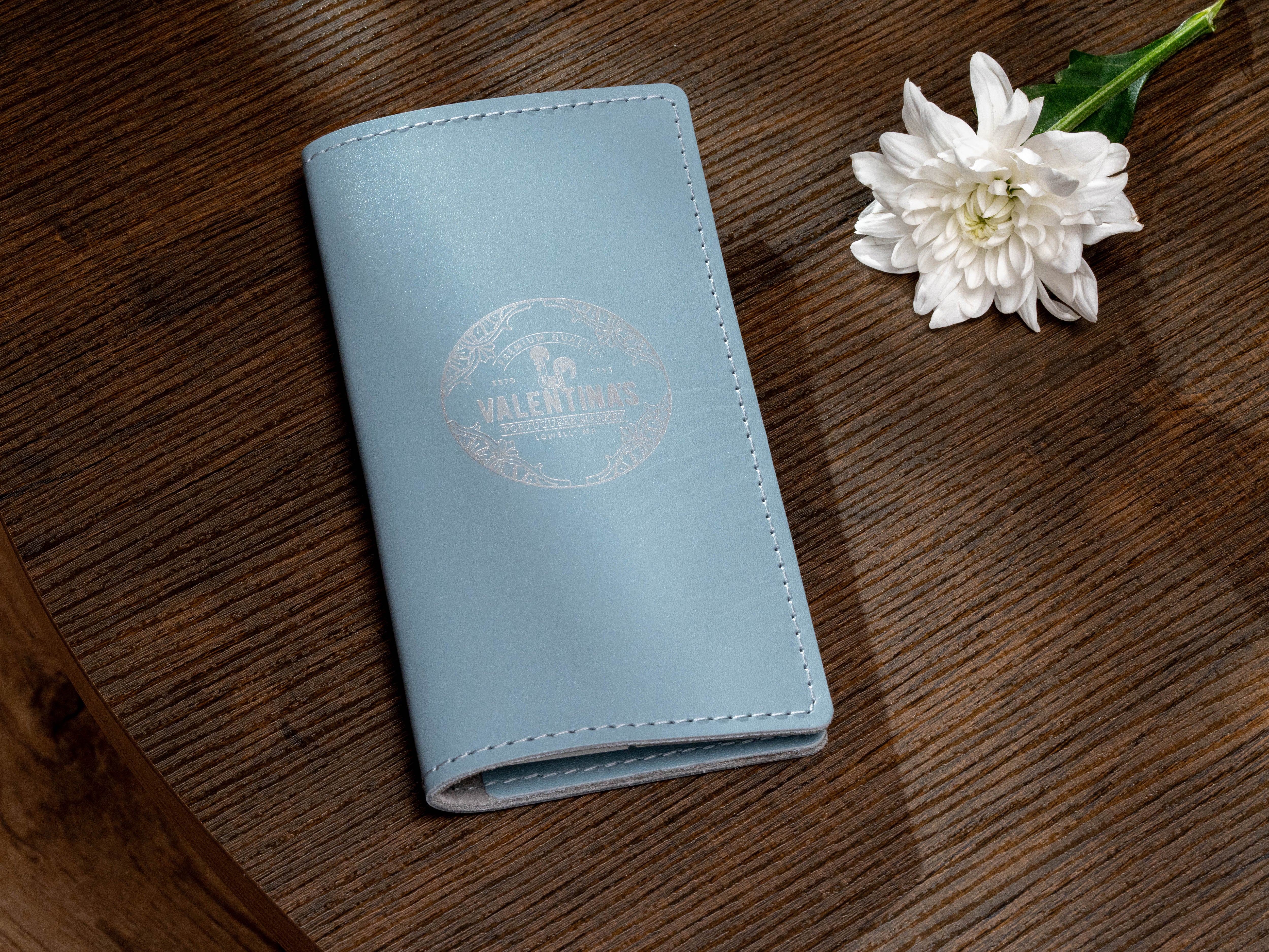 Ice blue leather receipt and payment folder with custom Portuguese market logo displayed beside white flower