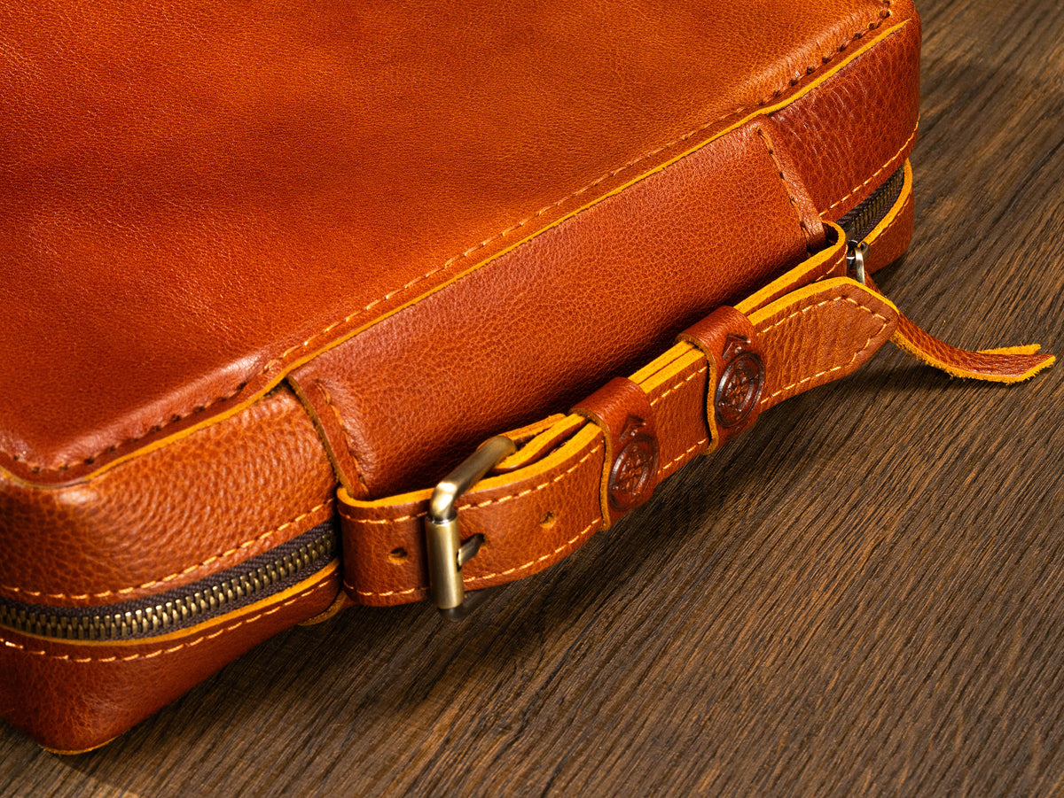Camel leather travel organizer case close-up with belt-style handle, compass details and hand saddle stitching
