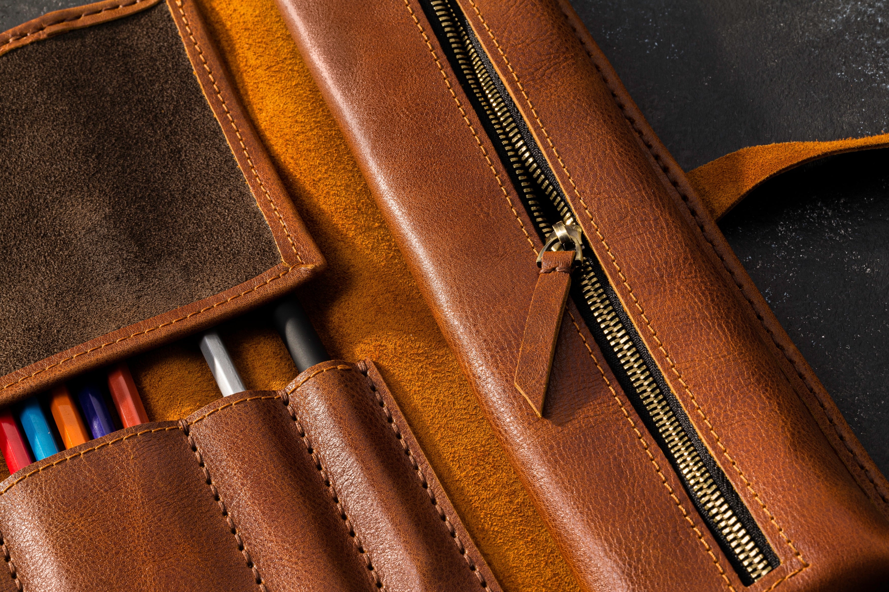 Brown leather pencil case with colorful pencils and a zipper on a dark surface