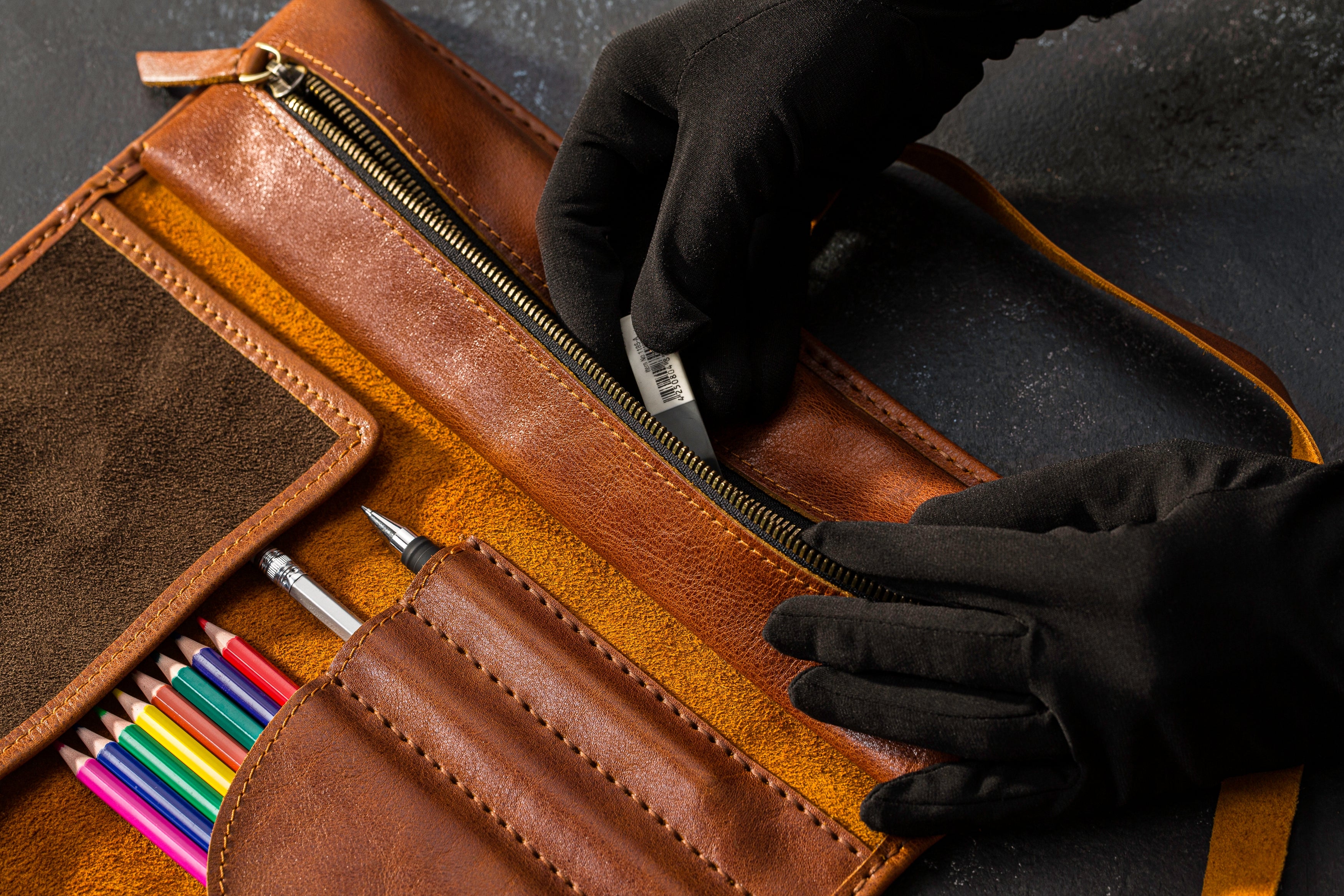 Brown leather case with colorful pencils and a hand wearing a black glove opening it.