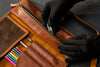 Brown leather case with colorful pencils and a hand wearing a black glove opening it.