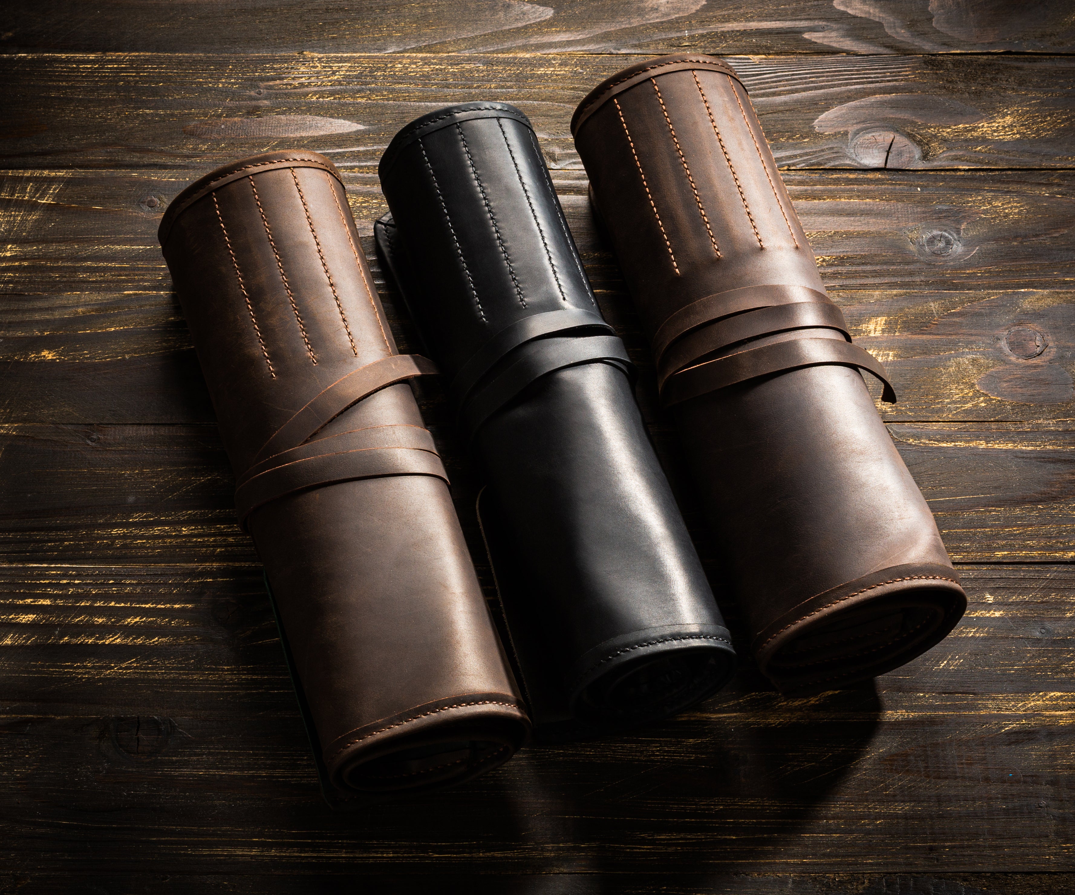 Trio of leather artist brush rolls in brown, black, and chestnut colors on wooden background